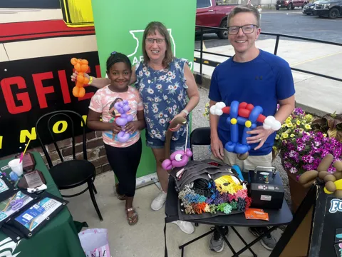 Two adults and a child pose beside colorful balloon creations and a display.