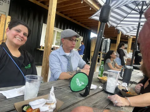 Participants sharing food and conversation at outdoor picnic tables. Alternate view 4.