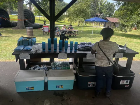 A participant stands beside a refreshment table under a shelter.