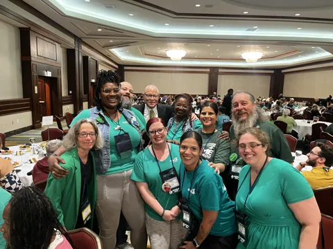 A group wearing green gathers for a photograph around a table in a meeting hall. Alternate view 2.