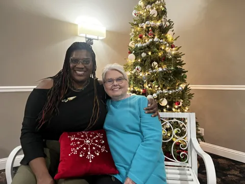 Two people pose beside a decorated Christmas tree. Alternate view 2.