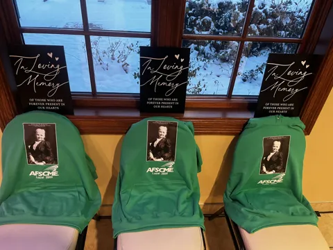 Three green AFSCME shirts and portrait photographs are displayed on chairs beneath memorial signs.