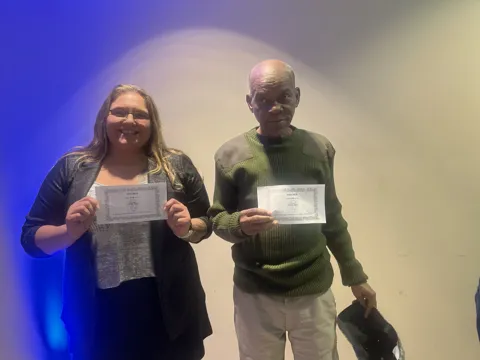 Two participants hold certificates at an indoor gathering.