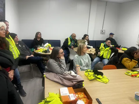 Participants gather in an office meeting space, some wearing high-visibility vests.