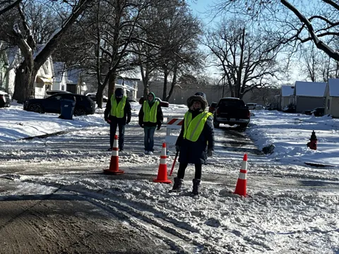 Participants wearing high-visibility vests stand on a snowy roadside near traffic cones. Alternate view 2.