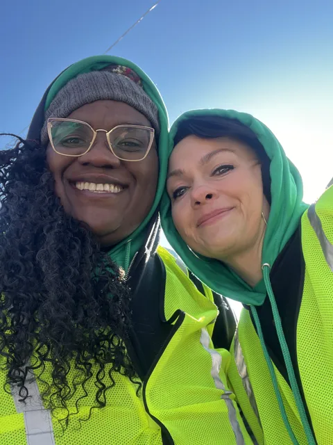 Two participants wearing high-visibility vests smile for a selfie in the snow.