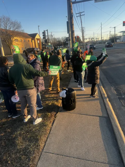 A group gathers on a sidewalk, with bags and belongings nearby. Alternate view 2.