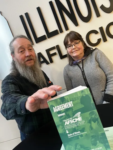 Two people pose together with an AFSCME agreement booklet beneath an Illinois AFL–CIO sign. Alternate view 3.