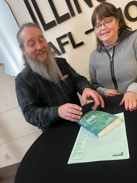 Two people pose together with an AFSCME agreement booklet beneath an Illinois AFL–CIO sign. Alternate view 4.