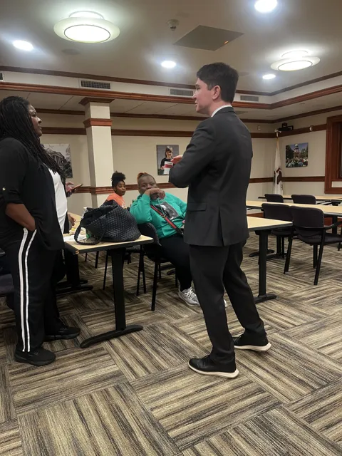 A speaker addresses participants seated around meeting tables.