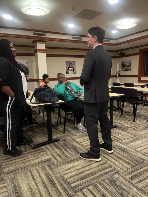 A speaker addresses participants seated around meeting tables. Alternate view 2.