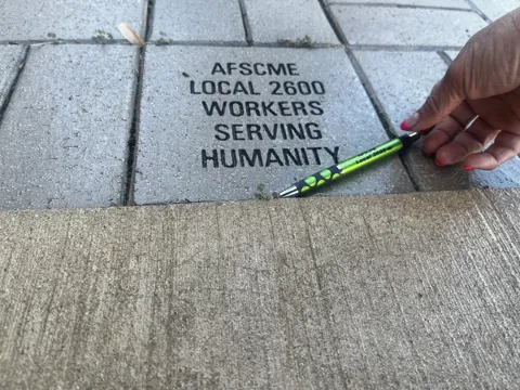 A hand places a pen beside a stone engraved “AFSCME Local 2600 Workers Serving Humanity.”