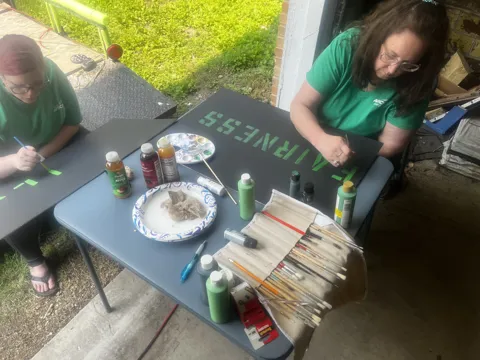Participants paint green lettering onto signs at an outdoor table.