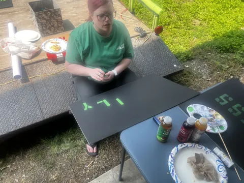 Participants paint green lettering onto signs at an outdoor table. Alternate view 4.